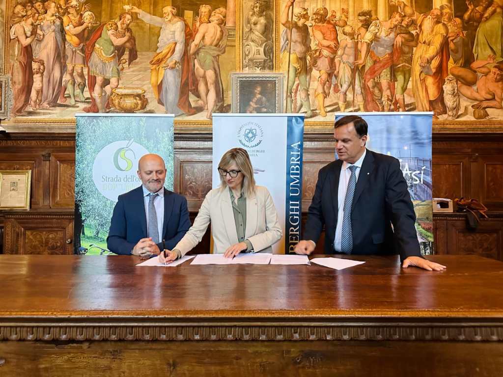Tre persone firmando un accordo di collaborazione per valorizzare l'olio extravergine di oliva, con banner di Strada dell'Olio e Federalberghi Umbria sullo sfondo, presso l'Abbazia di San Pietro a Perugia.