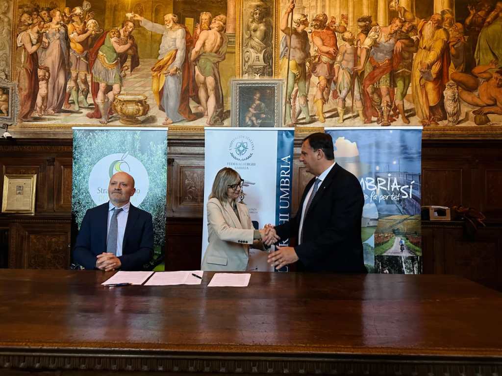 Due persone si stringono la mano durante la firma di un accordo di collaborazione, con opere d'arte sullo sfondo e cartelloni pubblicitari di promozione turistica dell'Umbria.