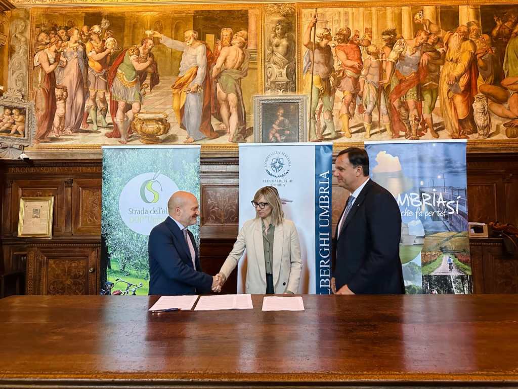 Tre rappresentanti di organizzazioni turistiche e agricole stringono la mano durante la firma di un accordo di collaborazione per promuovere l'olio extravergine di oliva come prodotto identitario dell'Umbria, con uno sfondo artisticamente ricco.
