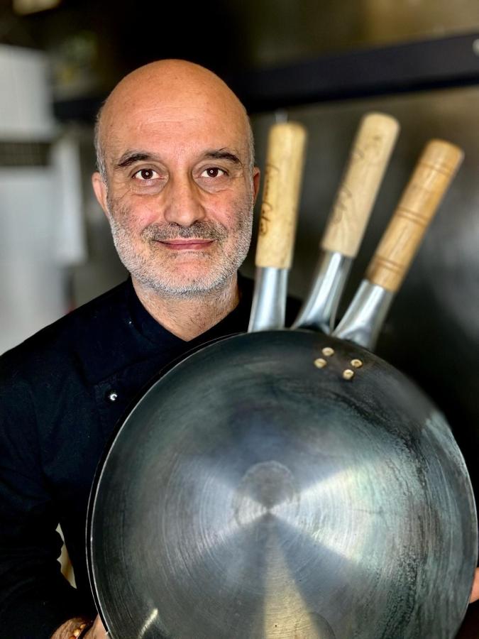 Chef Gianni Catani holding a large wok with three wooden-handled utensils in the background.
