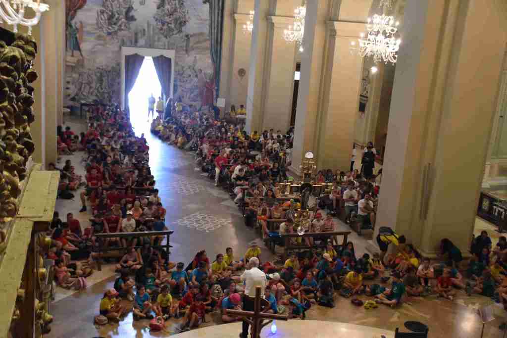 Gruppo di bambini e animatori riuniti in una cattedrale, ascoltando un intervento da un relatore con un grande numero di partecipanti colorati seduti a terra.