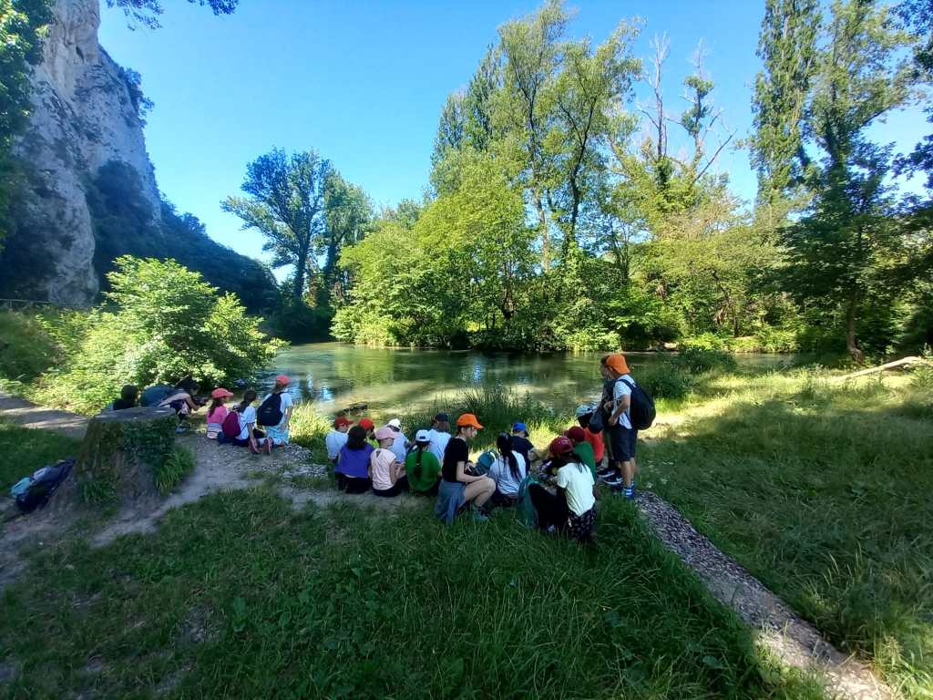Gruppo di bambini seduti vicino a un fiume, circondati da alberi e vegetazione lussureggiante, durante una gita nella natura.