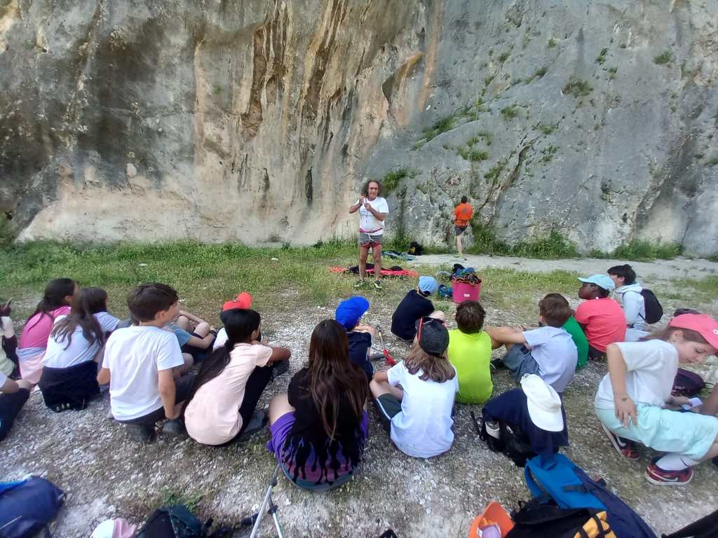 Gruppo di bambini seduti su un terreno erboso ascoltano un istruttore di arrampicata mentre spiega la tecnica, con una parete rocciosa sullo sfondo.