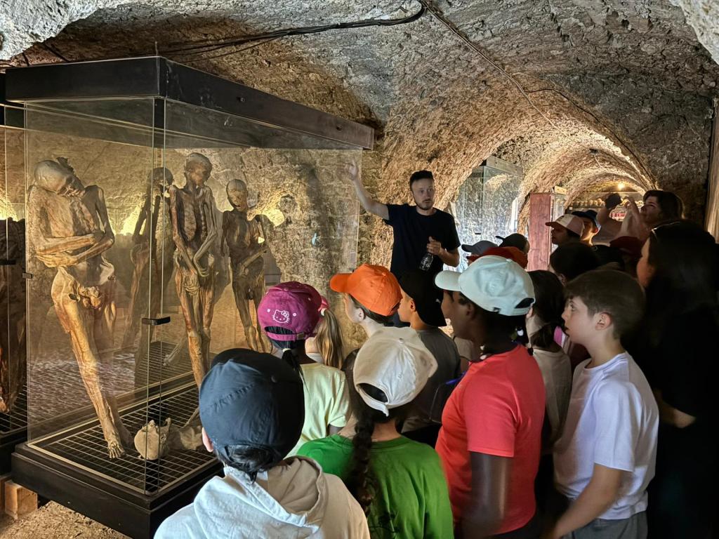 Bambini in visita al Museo delle Mummie di Ferentillo, ascoltando una guida che spiega la storia delle mummie, all'interno di un ambiente roccioso.