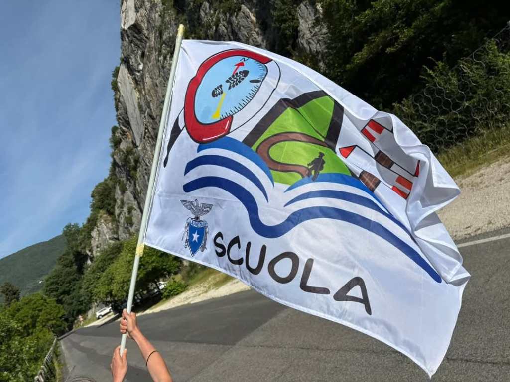 Una persona tiene in mano una bandiera con il logo del CAI Scuola, sullo sfondo si vedono alberi e scogliere.