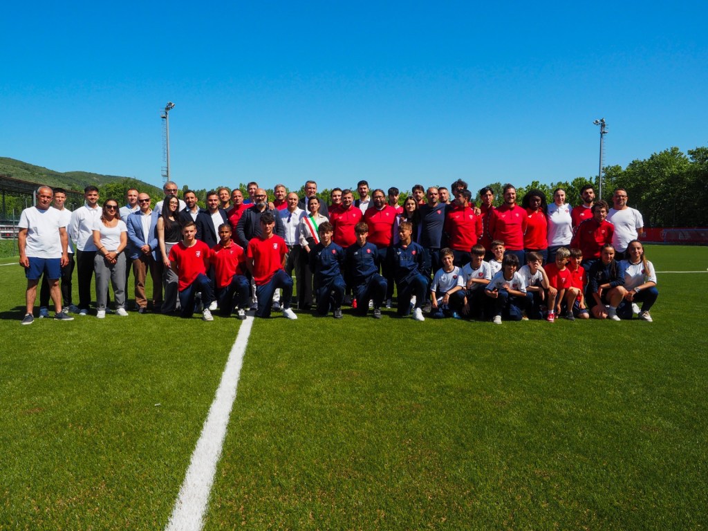 Gruppo di persone sorridenti in piedi su un campo da calcio sintetico appena inaugurato, con il cielo blu sullo sfondo.