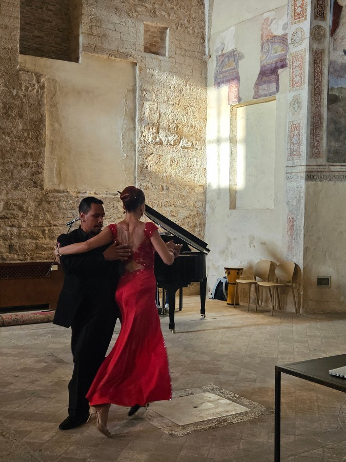 Due ballerini danzano insieme in una sala con pareti in pietra e affreschi, mentre un pianoforte è visibile sullo sfondo.