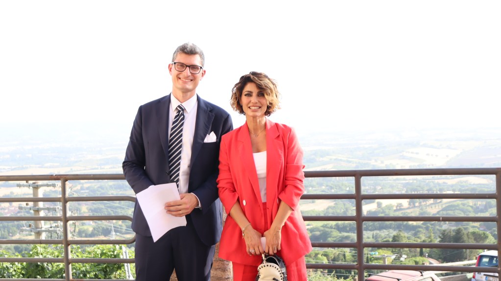 Due persone sorridenti in piedi su un balcone con vista panoramica. L'uomo indossa un abito scuro e una cravatta, mentre la donna è vestita con un completo rosso. Sullo sfondo si vede un paesaggio di colline.