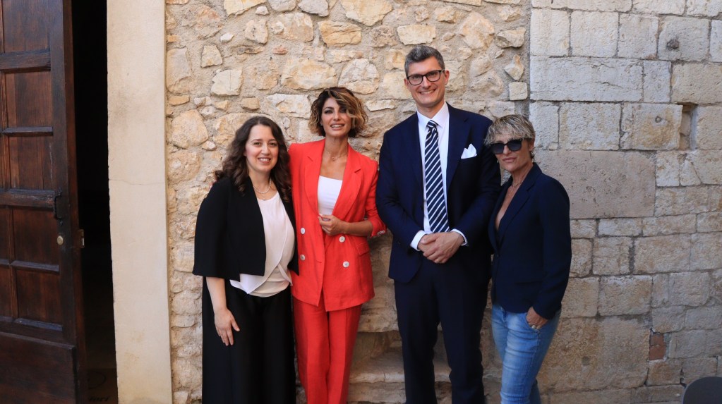 Quattro persone sorridenti posano davanti a una parete di pietra, due donne in abiti eleganti, un uomo in giacca e cravatta, e una donna con giacca blu e jeans.