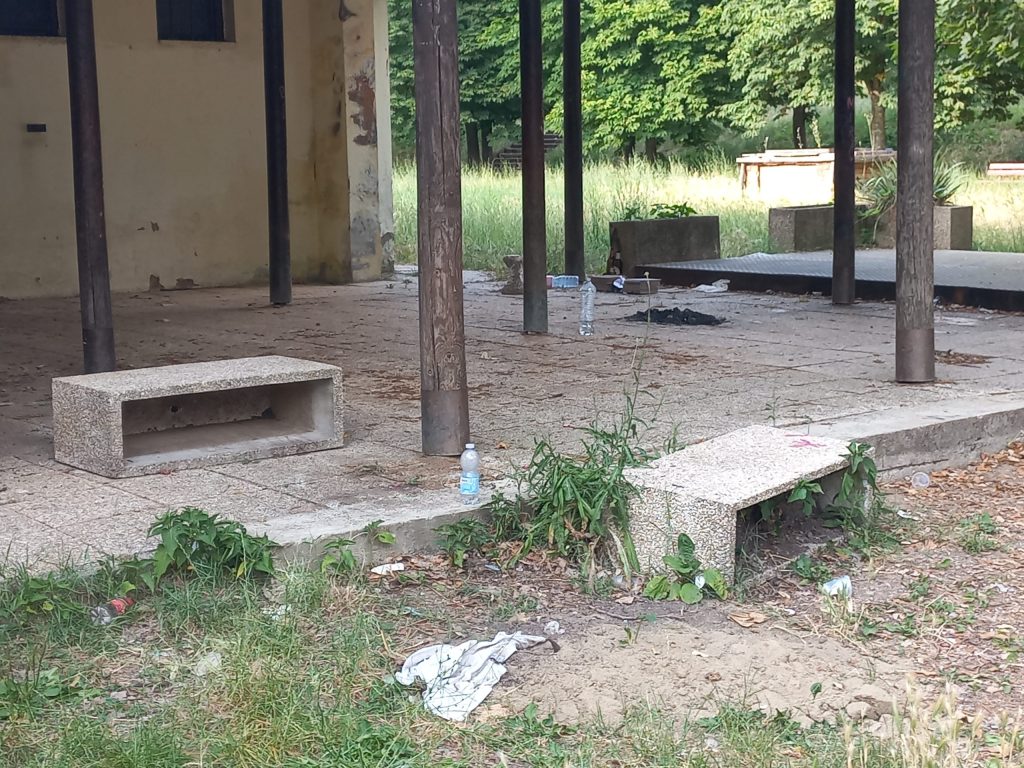 Area degradata del Parco dei Rimbocchi con erba alta, rifiuti e attrezzature abbandonate.