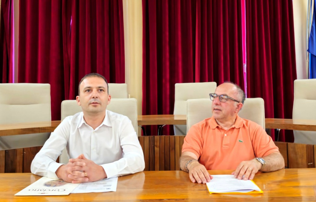 Due uomini seduti a una tavola in una sala, con tende rosse sullo sfondo. Uno indossa una camicia bianca, l'altro una maglietta arancione.