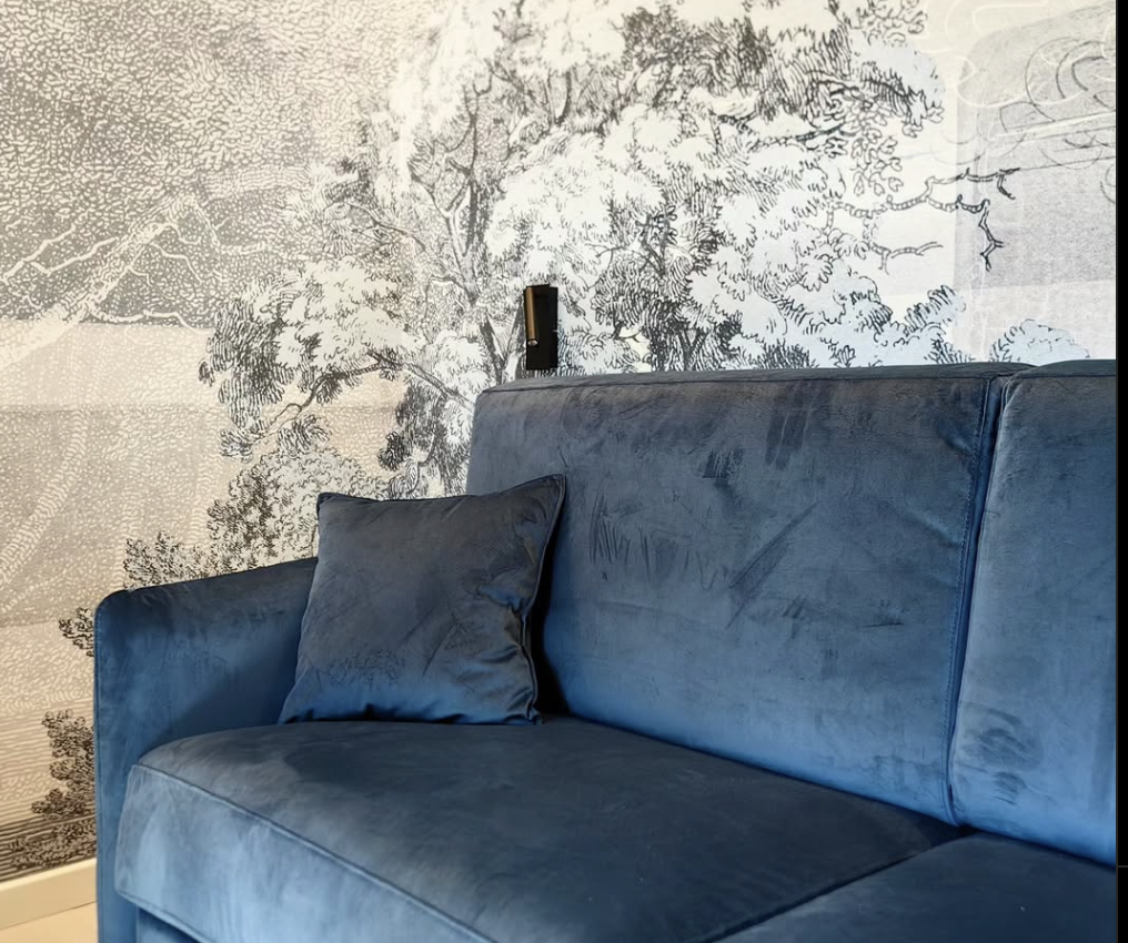 Un divano blu opaco con un cuscino sullo sfondo di una carta da parati artistica che rappresenta un paesaggio di alberi.