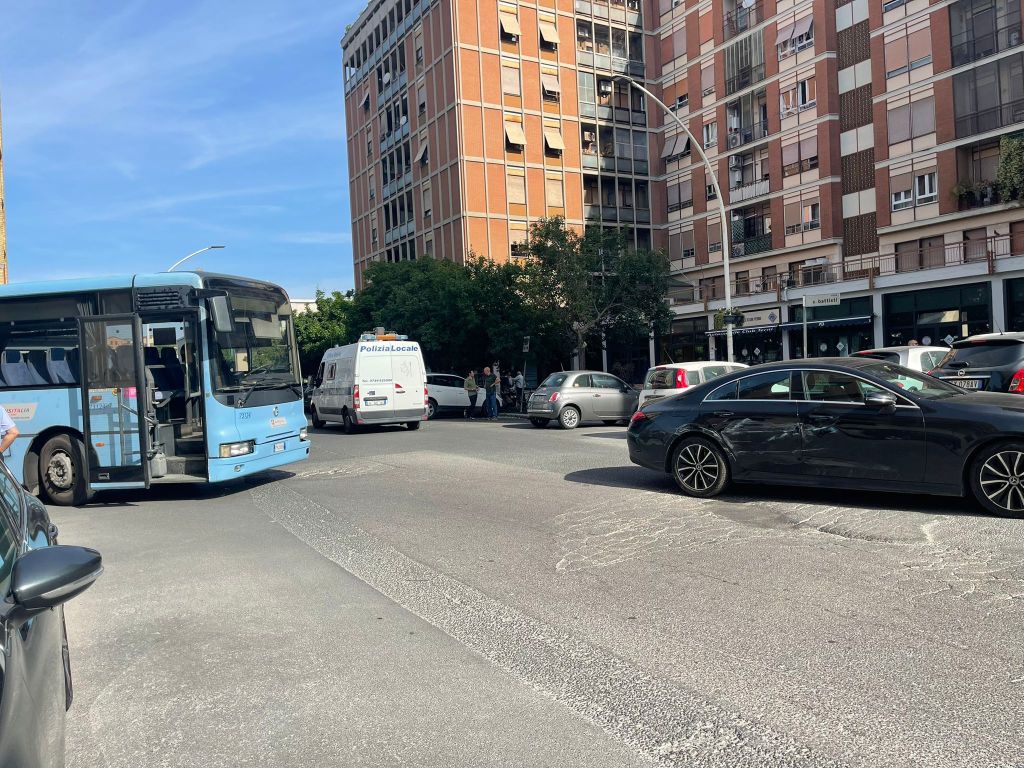 Una scena di un incidente stradale tra un autobus blu e una berlina nera in una strada ben illuminata, con edifici residenziali sullo sfondo e la polizia locale in arrivo.