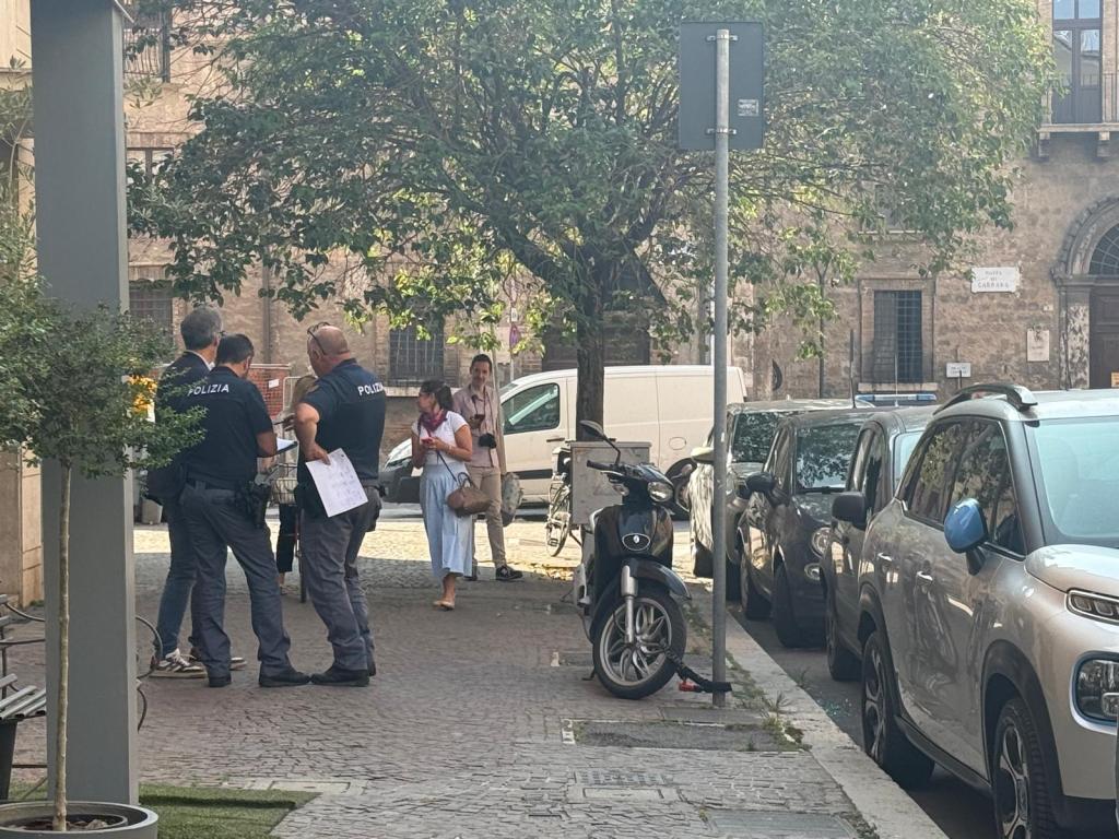 Un gruppo di agenti di polizia discute in strada mentre una donna osserva. Gli agenti indossano abbigliamento della polizia e tengono documenti. Sullo sfondo, ci sono auto parcheggiate e un ambiente urbano.