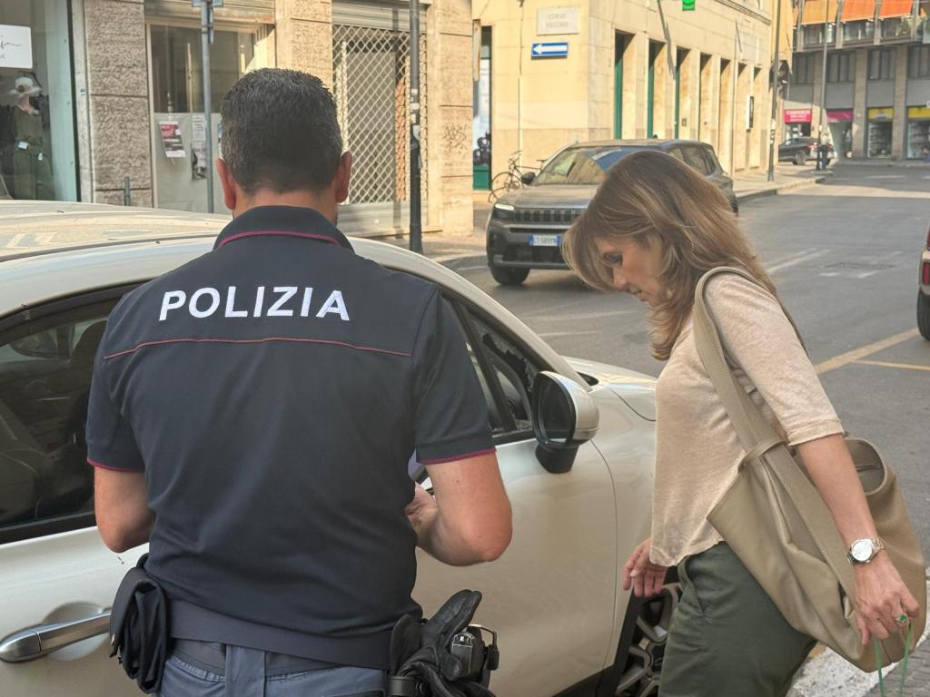 Un agente di polizia con la divisa leggermente visibile controlla un'auto mentre una donna, con una borsa a spalla, guarda verso il veicolo.