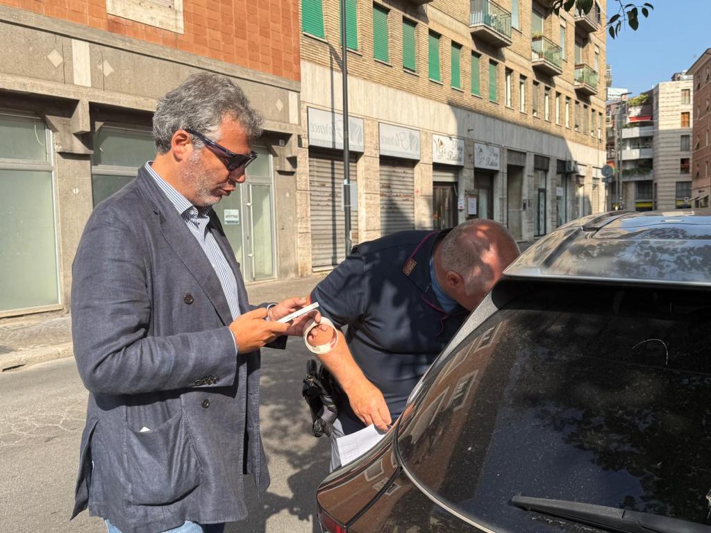 Due uomini discutono accanto a un'auto danneggiata in una strada di Terni, con un edificio a più piani sullo sfondo.