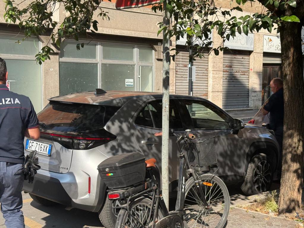 Un agente di polizia esamina un'auto danneggiata in una strada di Terni, con una bicicletta parcheggiata accanto, mentre si intravede un negozio chiuso sullo sfondo.