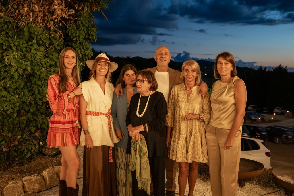 Un gruppo di sette persone sorridenti posano insieme all'esterno al crepuscolo, con vegetazione e automobili sullo sfondo.