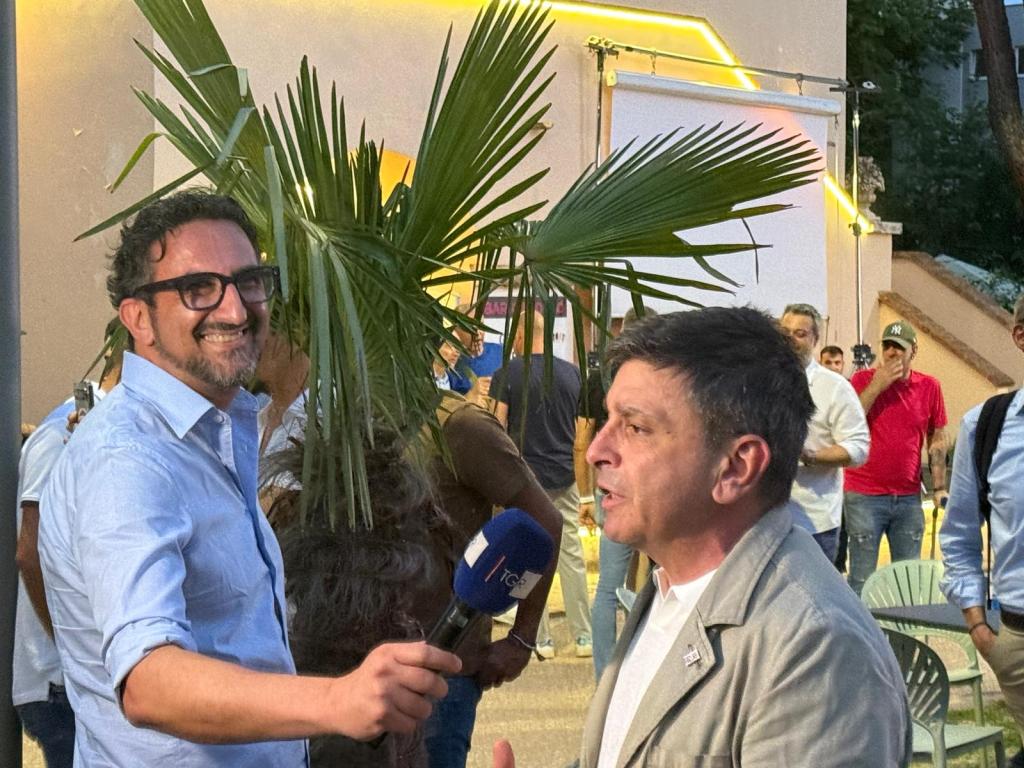 Due uomini in un evento all'aperto, uno dei quali sta facendo un'intervista mentre un pubblico si riunisce dietro di loro. Sullo sfondo si vedono alberi e decorazioni luminose.