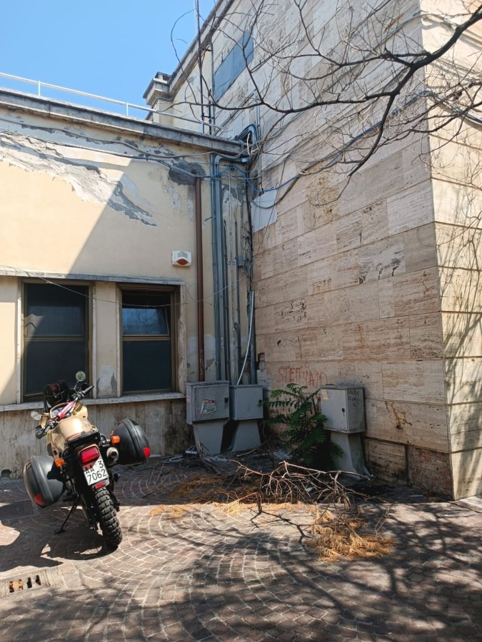 An abandoned corner of a building with peeling paint and exposed pipes, featuring a motorcycle parked in front. The ground is made of cobblestones, and there are scattered branches and debris.