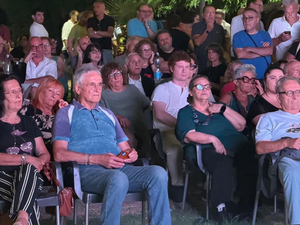 Un gruppo di persone sedute durante un evento all'aperto, osservando attentamente di fronte a loro, con illuminazione soffusa e atmosfera serale.