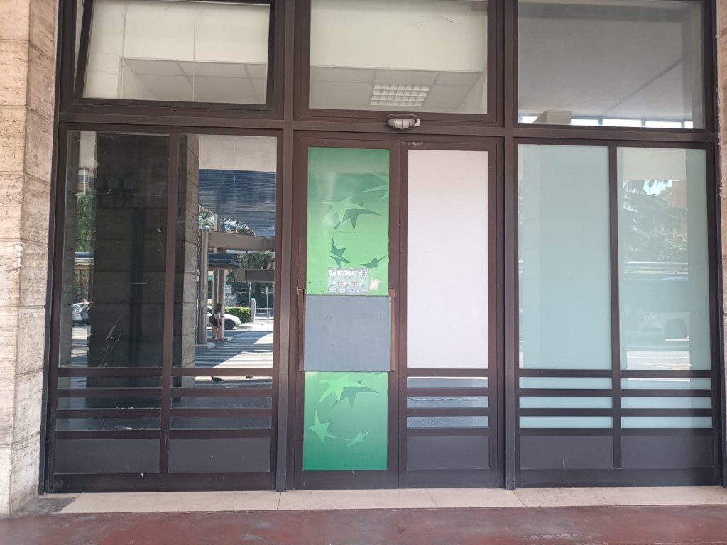 Front view of a commercial building entrance with large glass windows and a green promotional poster displayed.