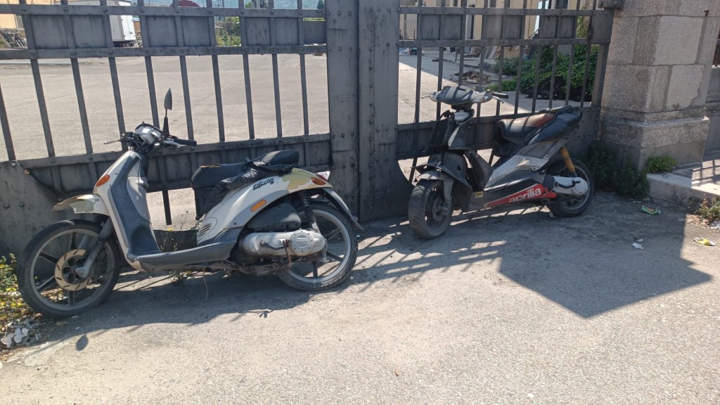 Two scooters parked beside a metal gate on a sunny day.