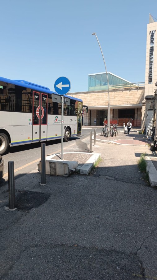 A city bus stops at an intersection near a building, with bicycles parked nearby and a road sign indicating a left turn.
