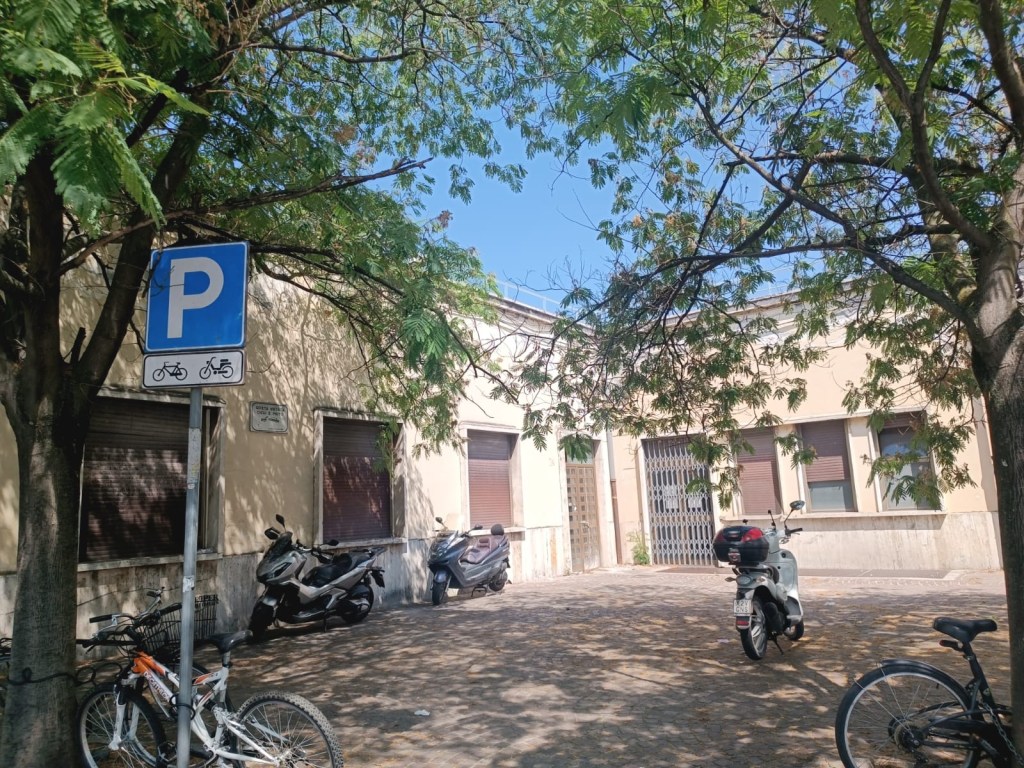 A parking area with a blue 'P' sign for bicycles and scooters located near a building. Nearby are several parked bicycles and scooters, with trees providing shade.