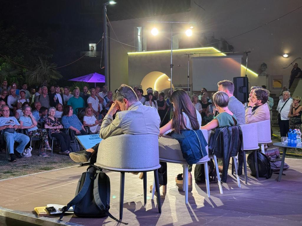 Un incontro pubblico con un gruppo di relatori seduti su un palco, di fronte a un'ampia folla di cittadini in un evento all'aperto, con illuminazione serale.
