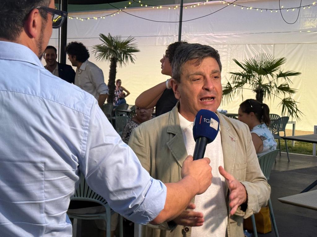 A man with short hair is speaking passionately into a microphone held by another person. The background features a tent-like structure with people seated and some greenery.