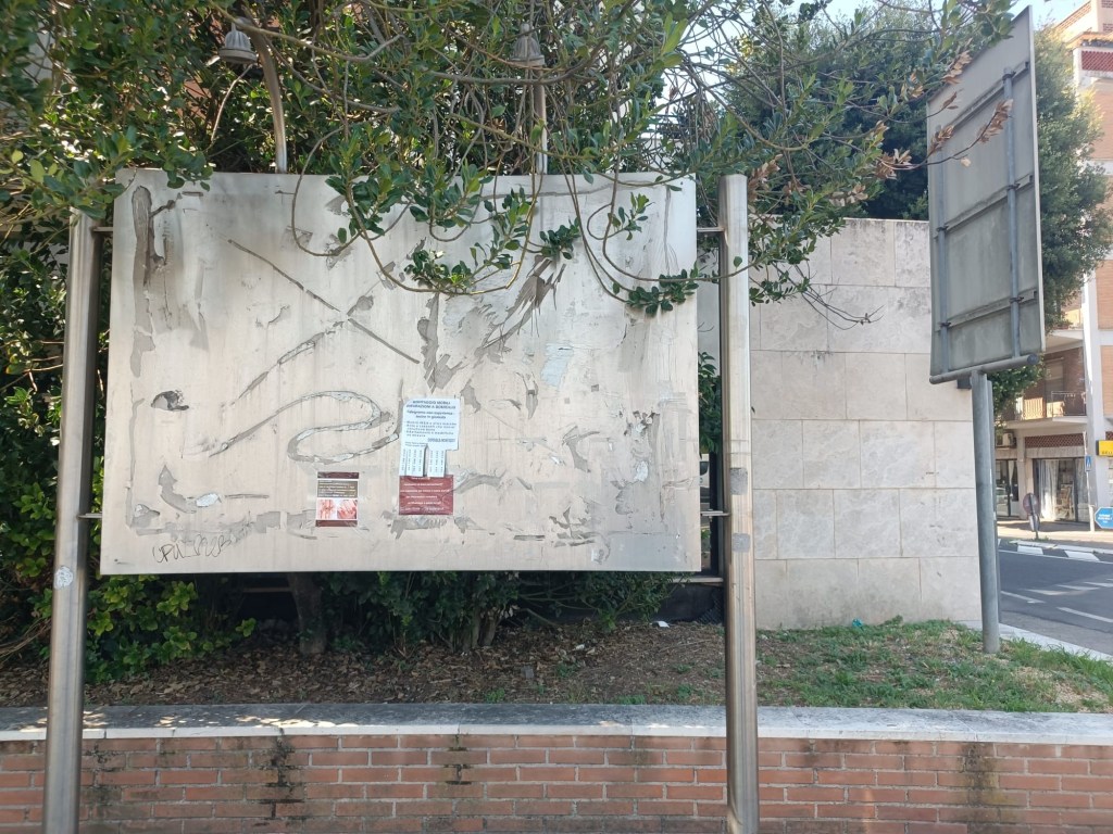 A large, empty billboard partially covered by tree branches and showing signs of wear and tear, located in an urban area.