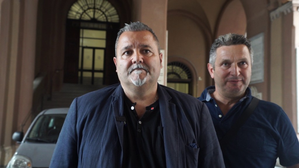 Due uomini in una hall, uno di fronte all'altro, con uno sfondo di porte e scale. Entrambi sorridono, mentre un'auto è visibile sul lato sinistro.