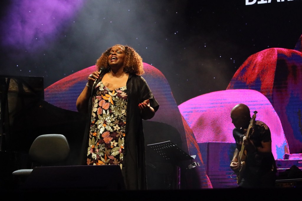 A female singer performs on stage, dressed in a floral outfit, with a guitarist playing beside her. Colorful lighting and abstract visuals create a vibrant atmosphere.