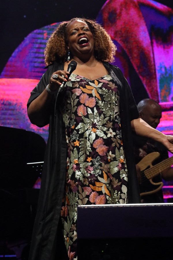 A female singer with curly hair joyfully performing on stage, holding a microphone, dressed in a colorful floral-patterned dress and black cardigan.