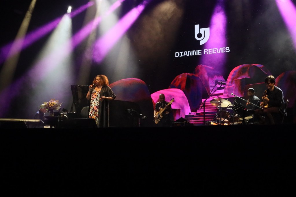 Jazz singer Dianne Reeves performing on stage with a band, illuminated by colorful stage lights.