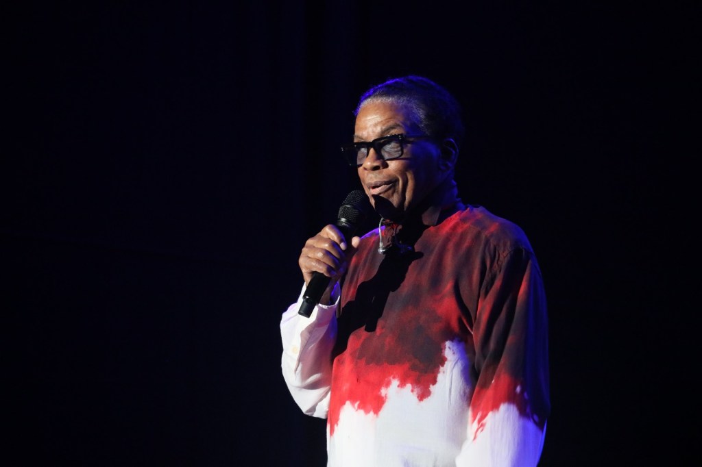 A person wearing glasses and a colorful shirt holding a microphone on stage, speaking in front of a dark background.