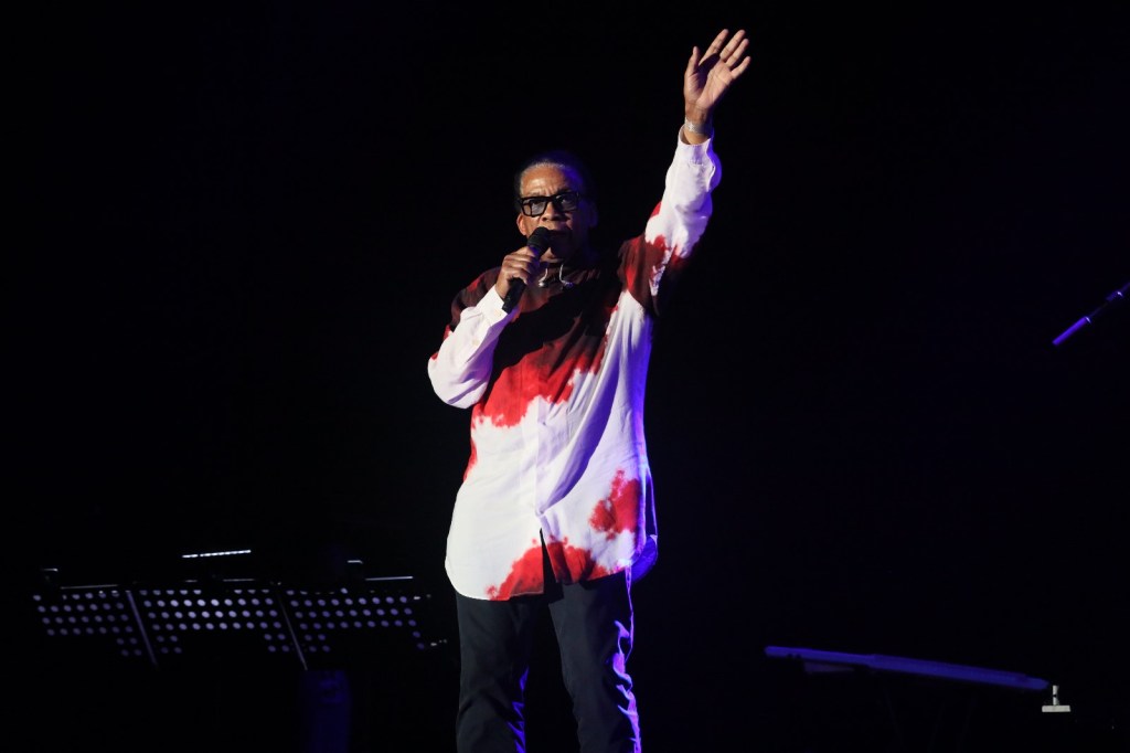 A performer on stage wearing a tie-dye shirt with red accents, passionately holding a microphone and gesturing with one hand.