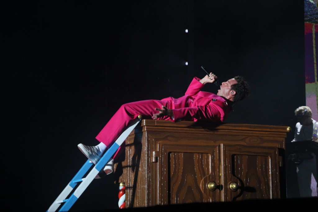 A singer in a bright pink suit performs on top of a wooden cabinet, reclining while holding a microphone, with a ladder leaning against it in a dramatic stage setting.