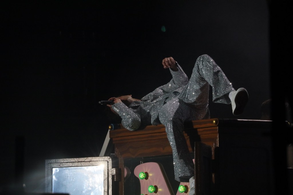 A performer in a sparkling silver suit lying on a wooden structure with a microphone in hand, illuminated by stage lights.