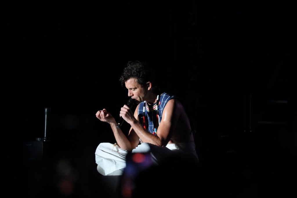 A solo artist performing on stage, holding a microphone and sitting on the floor, dressed in a sleeveless top and white pants, with a focused expression.