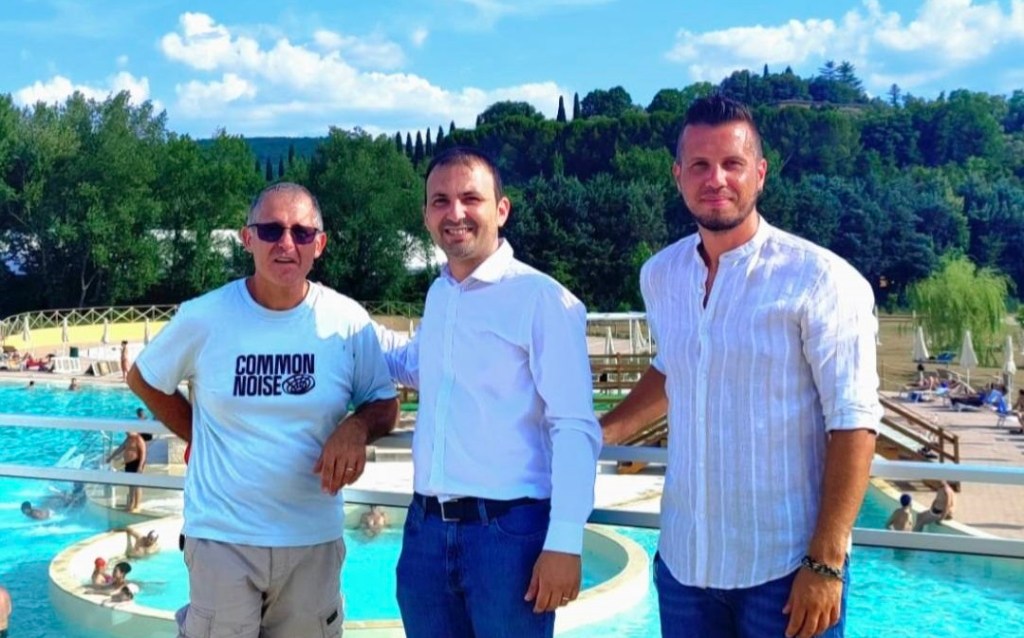 Tre uomini sorridenti in posa davanti a una piscina, con alberi e un cielo blu sullo sfondo.
