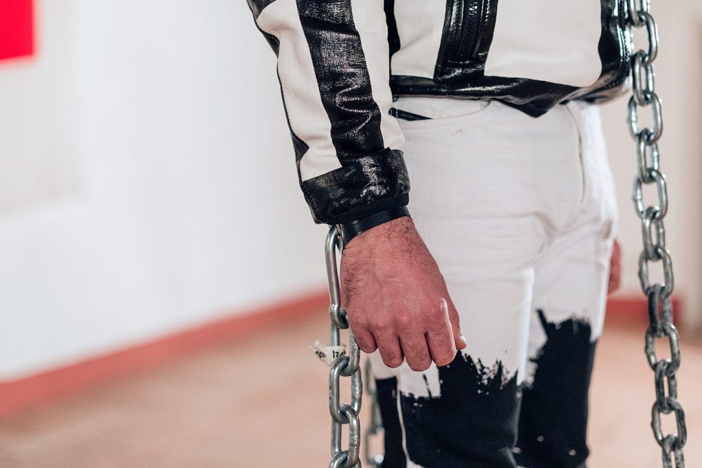 Dettaglio di una persona in abbigliamento bicolore, con una mano che tiene una catena. Il contesto sembra essere un ambiente artistico.