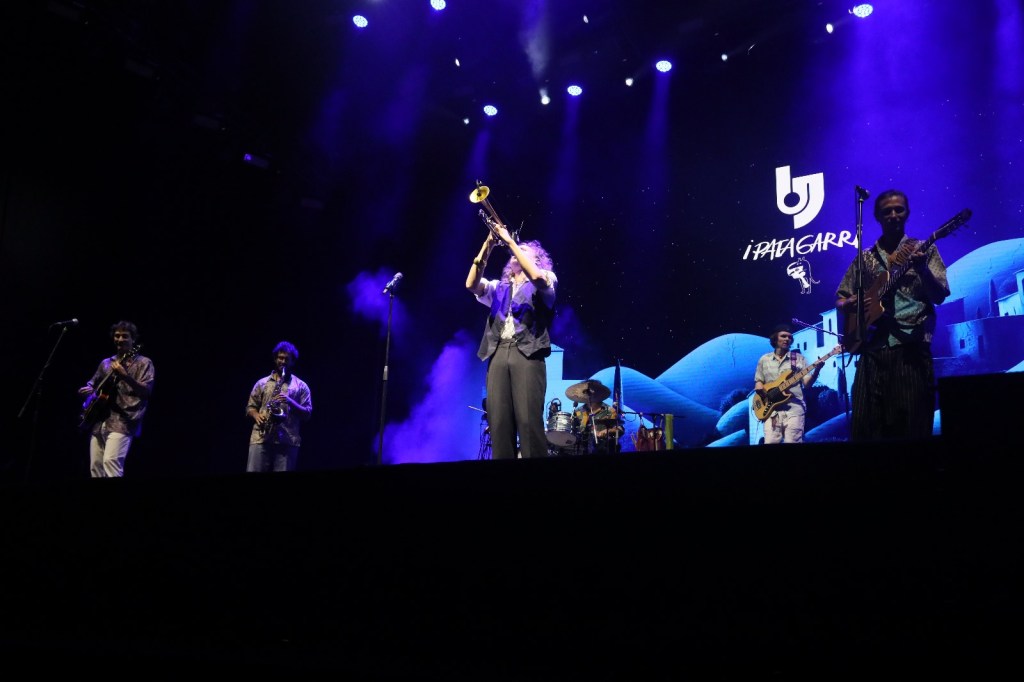 Musicians performing on stage with colorful lighting, featuring a trumpet player and a band set against a backdrop with a stylized logo.