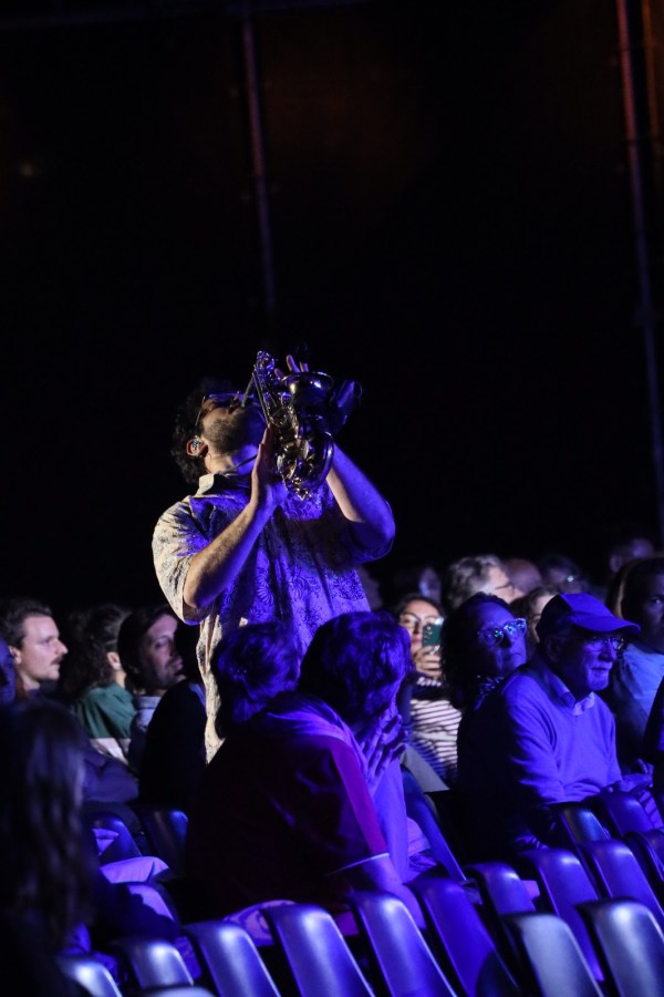 A musician playing a saxophone on stage with an audience in a dimly lit venue.