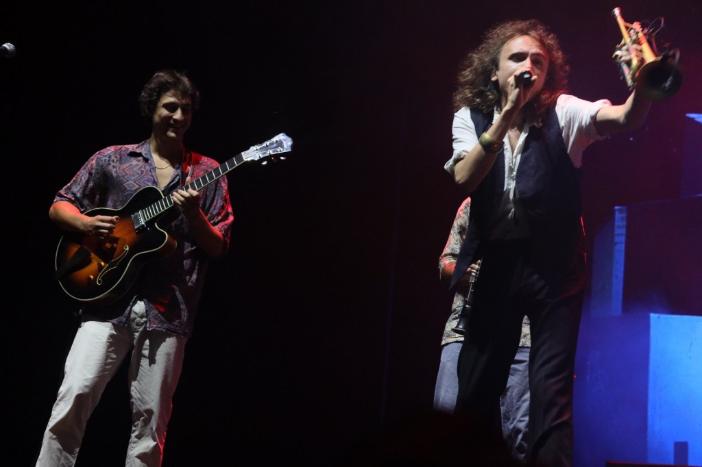 Un artista con una trompeta en el escenario mientras otro músico toca la guitarra, iluminados por luces de fondo en un concierto.