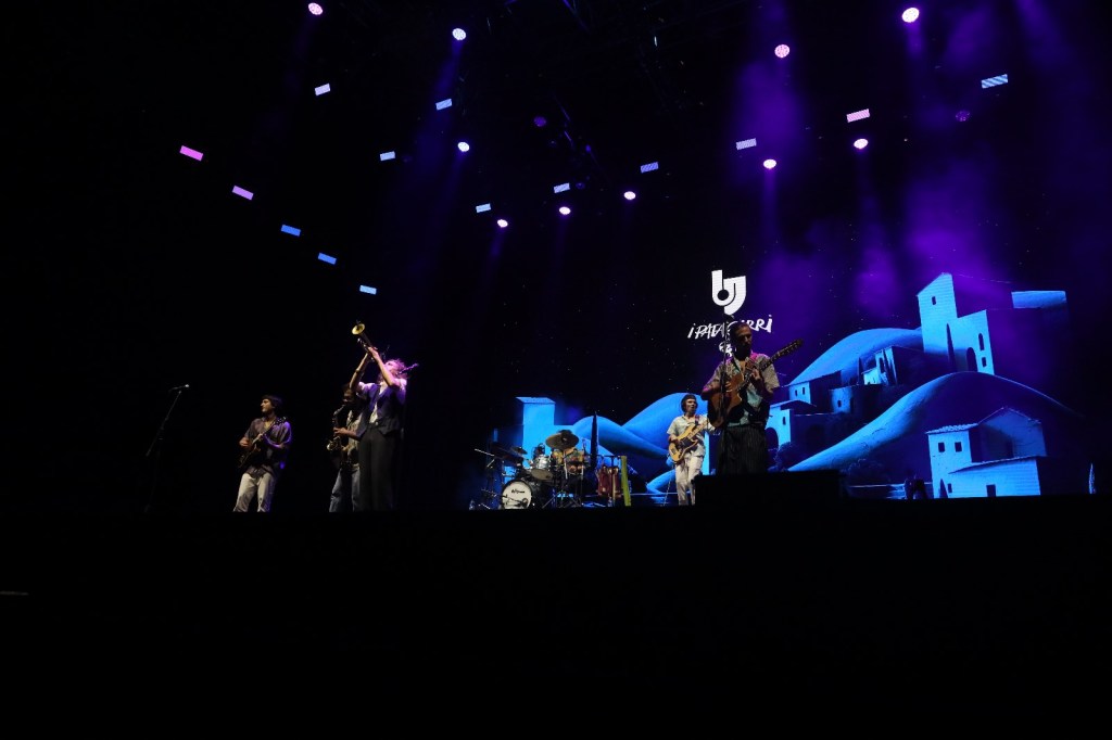 A live musical performance featuring a group of musicians on stage, illuminated by colorful lights, with a backdrop displaying stylized blue hills and houses.