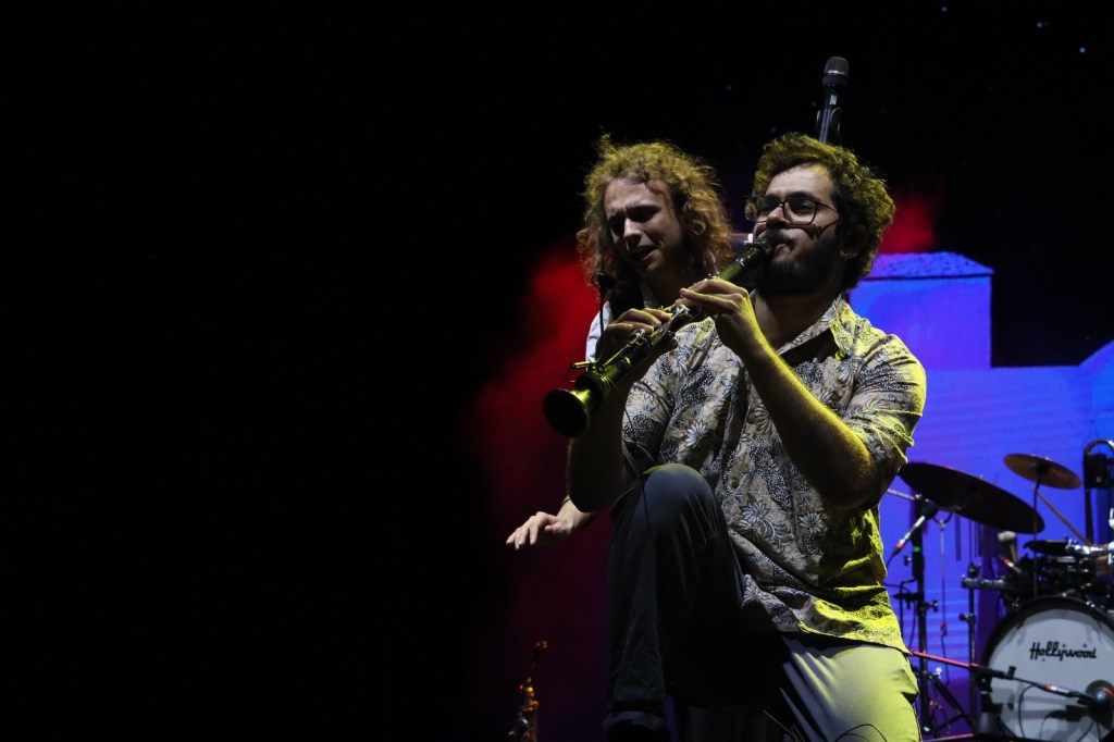 A musician performing on stage, playing a clarinet, with a lively background. Another person is seen in the back, adding to the energetic atmosphere.