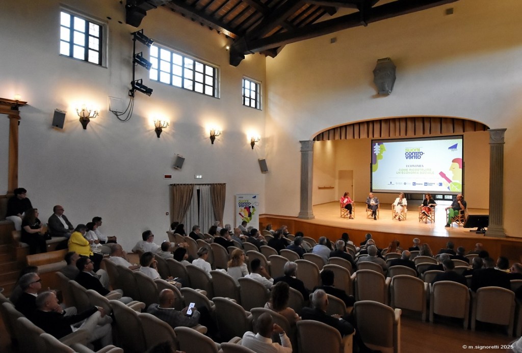 Un pubblico numeroso assiste a un incontro su economia sociale al Teatro Cucinelli di Solomeo, con relatori sul palco e proiezioni sullo sfondo.