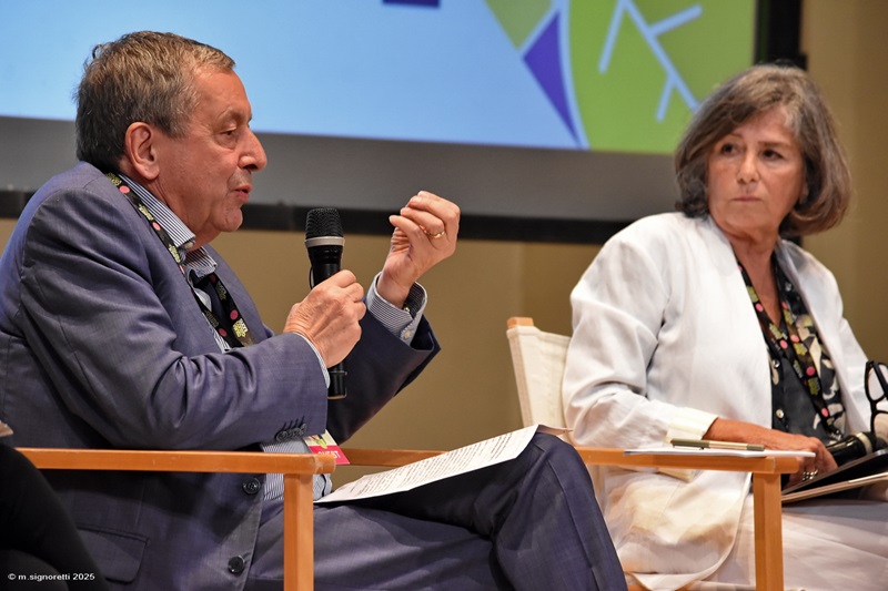 Panel discussion with two speakers, one gesturing while speaking into a microphone, and the other listening attentively, set in a conference environment.